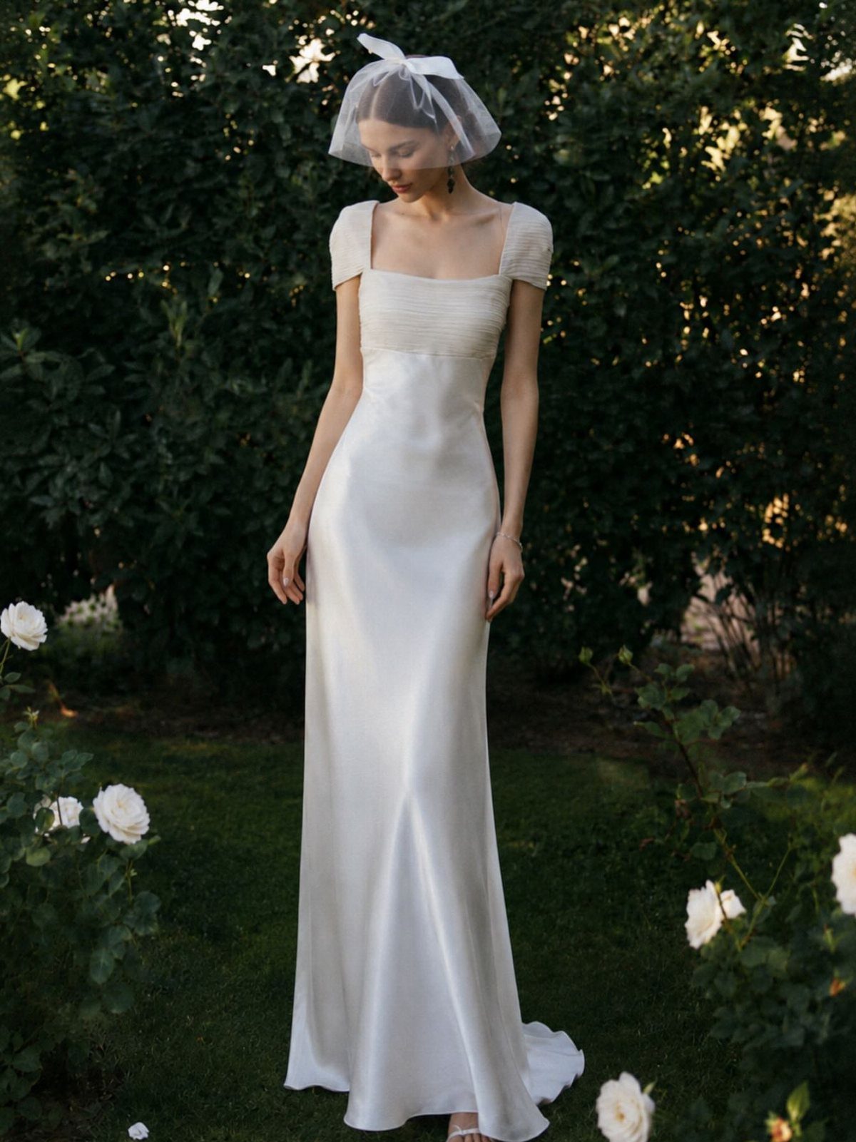 Woman in a sleek white silk wedding gown and birdcage veil in a garden