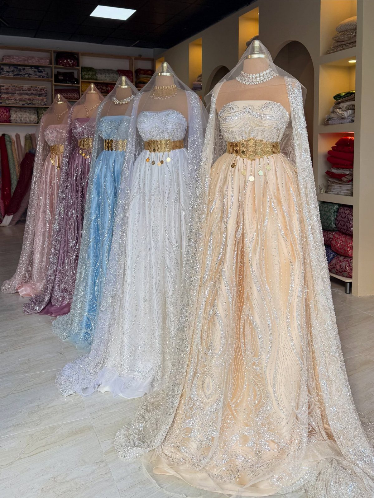 Five shimmering A-line gowns on mannequins featuring wide metallic gold bridal belts.