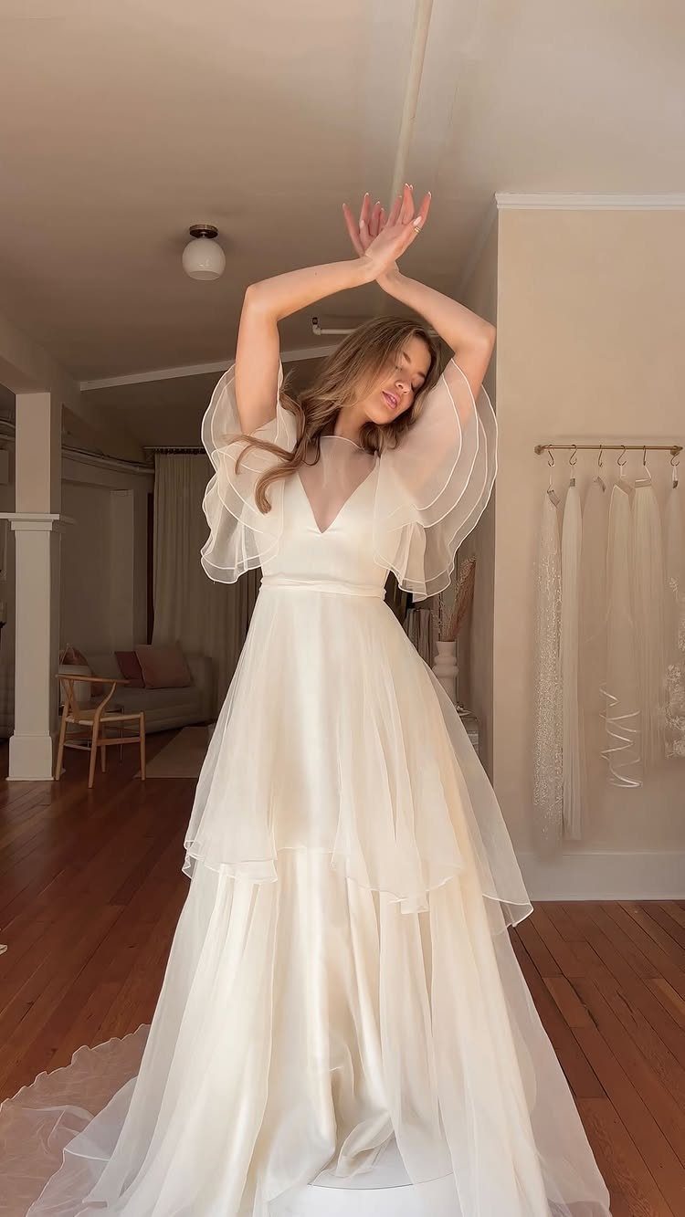 Woman in a white A-line wedding gown posing with arms raised indoors.