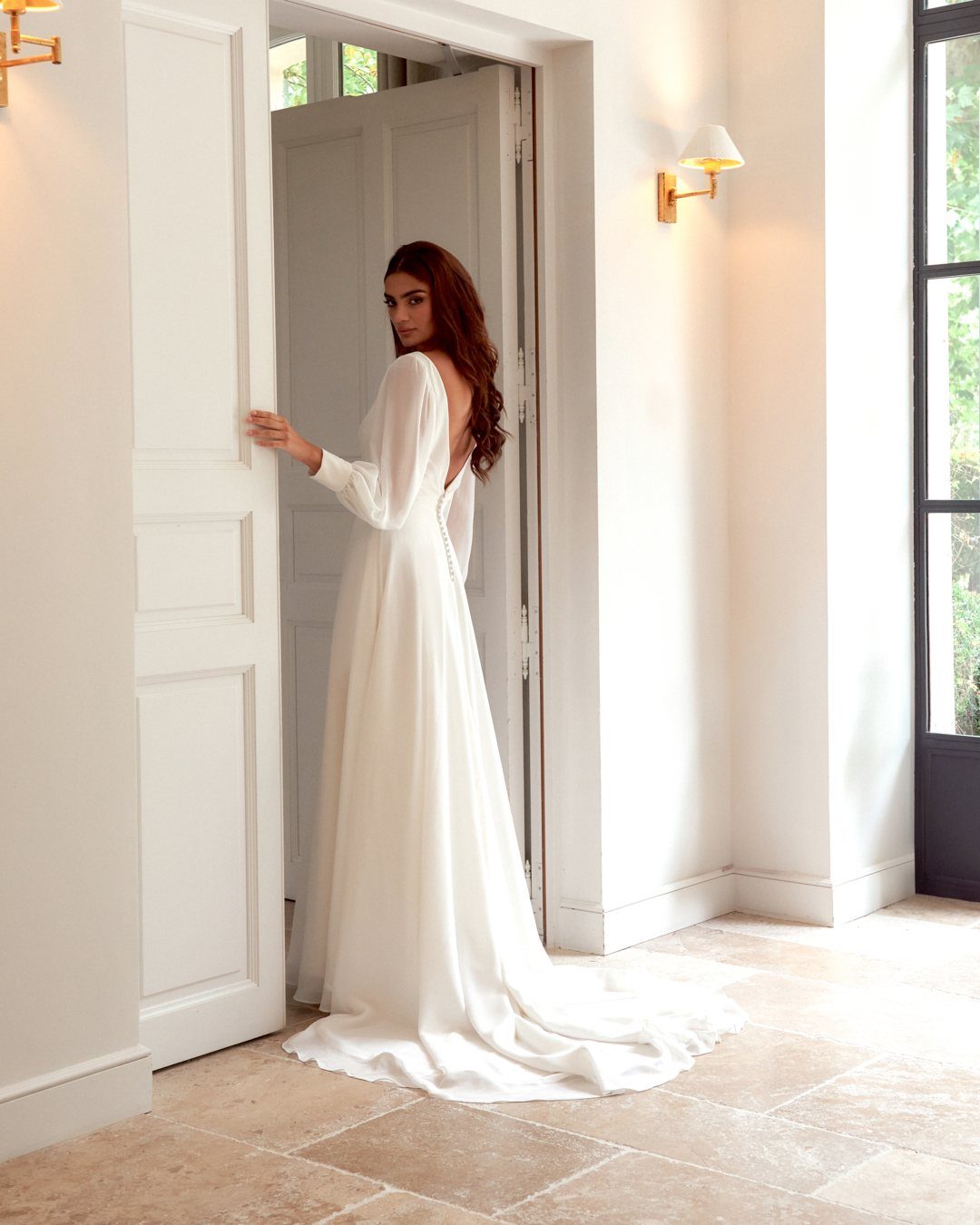 Woman in a white long-sleeved A-line wedding gown standing in a white doorway