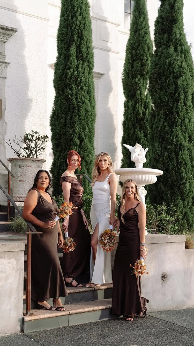 Three bridesmaids in dark brown dresses and a bride in white standing outdoors.