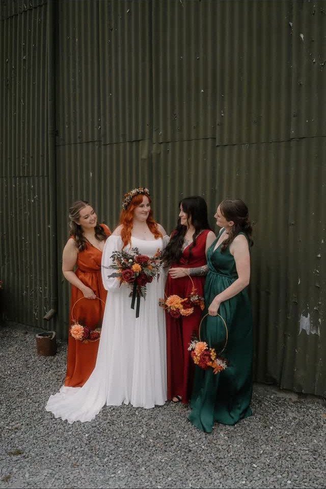 Bride and bridesmaids in jewel-toned autumn dresses posing against a green metal wall.