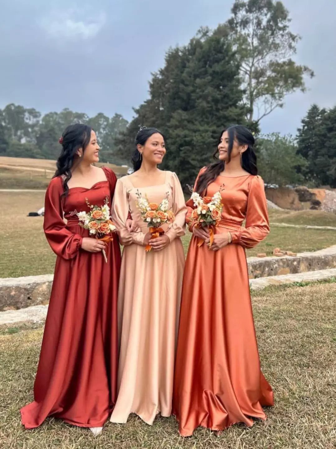 Three bridesmaids in long-sleeved satin dresses holding autumn bouquets in a field.