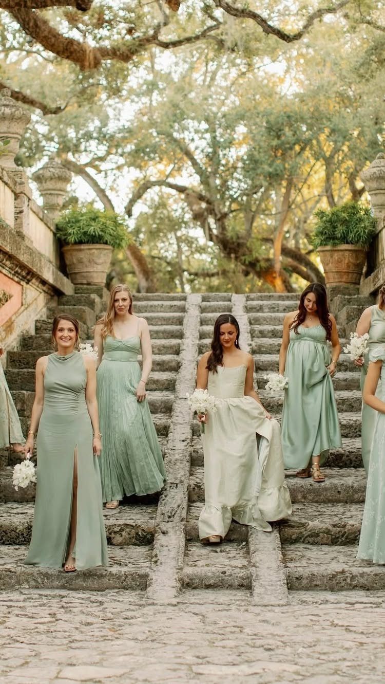 Bridesmaids in mismatched sage and pale green dresses standing on stone stairs.