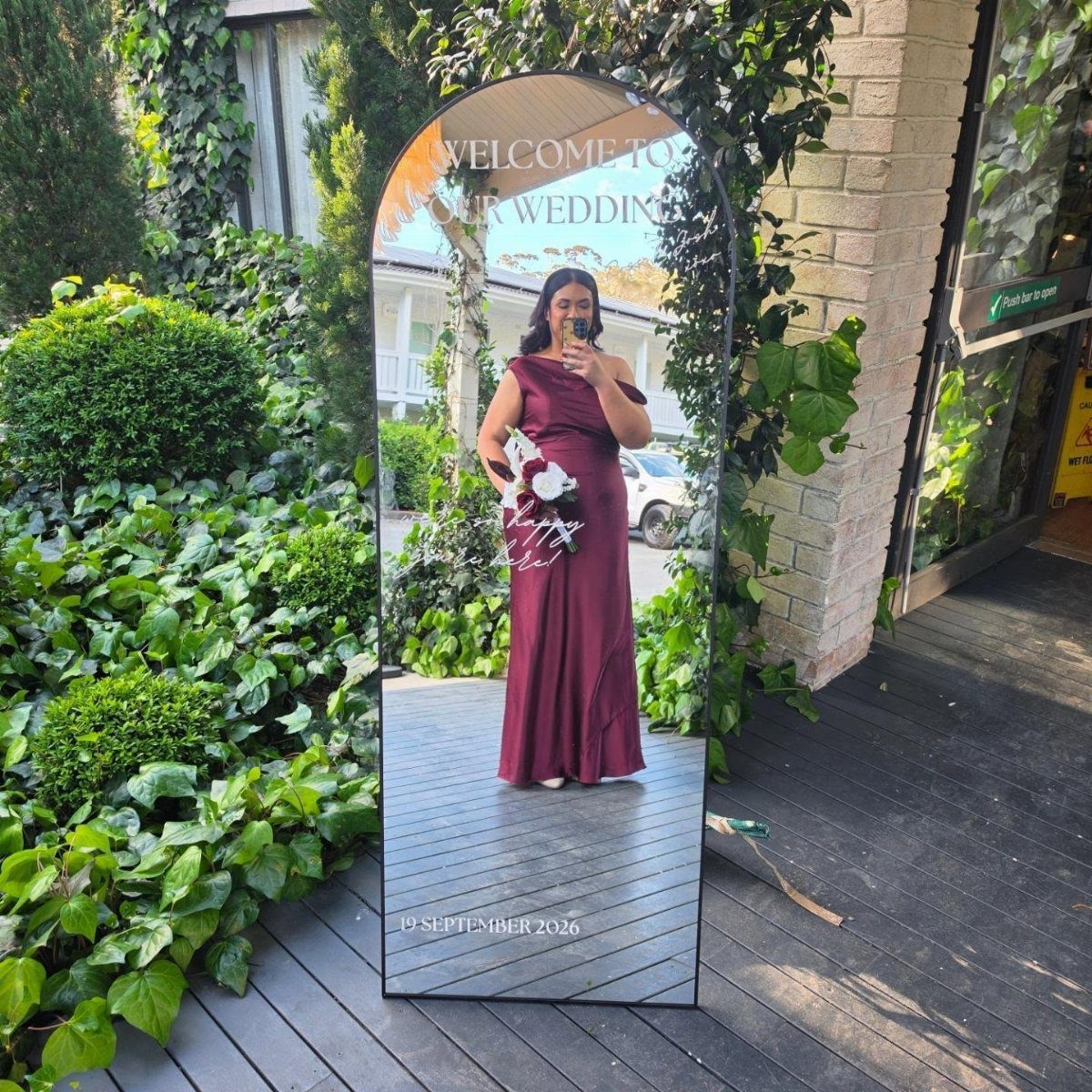 Woman in burgundy satin bridesmaid dress taking a mirror selfie outdoors with greenery.