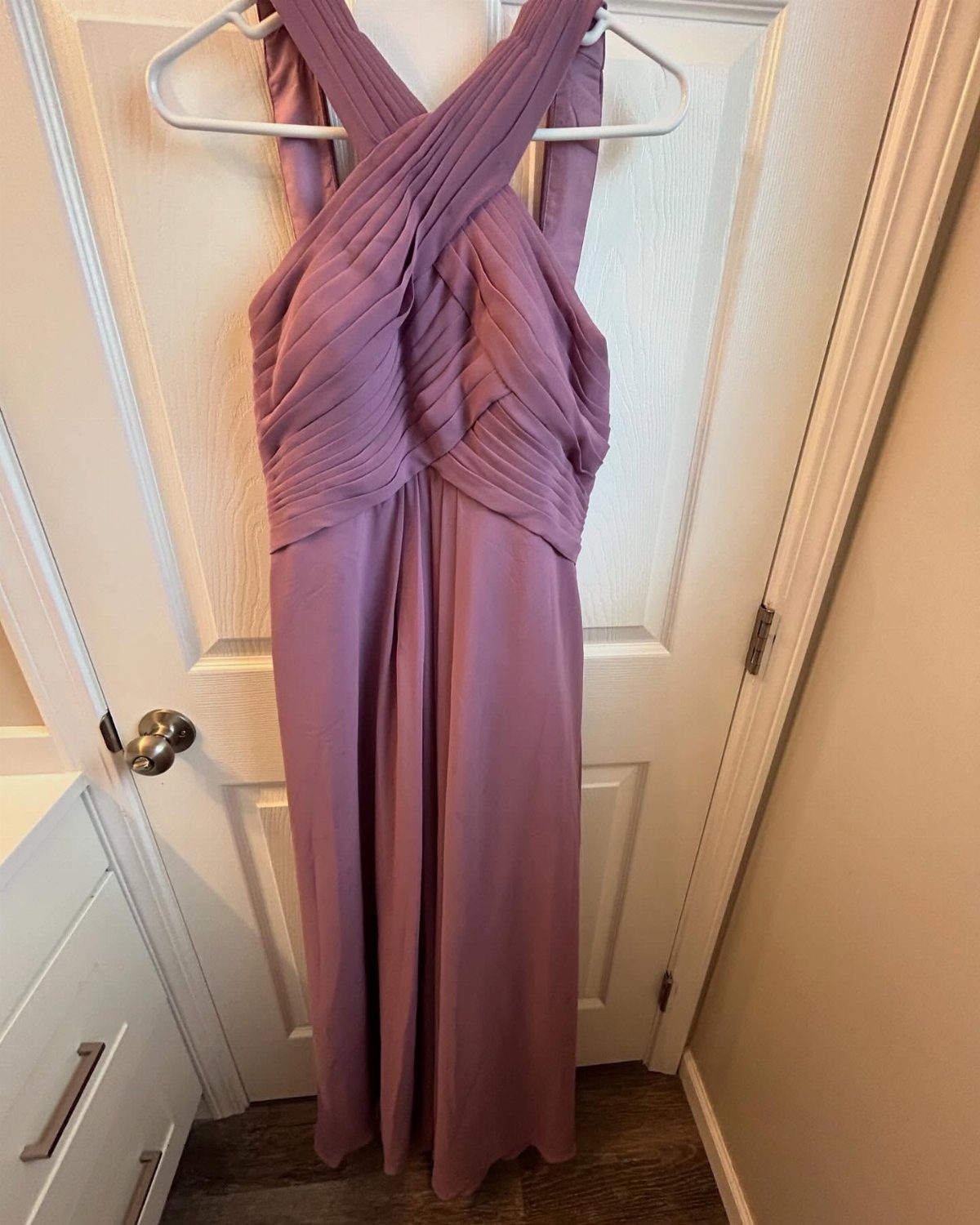 Mauve floor-length bridesmaid dress with pleated criss-cross bodice hanging on a white door.