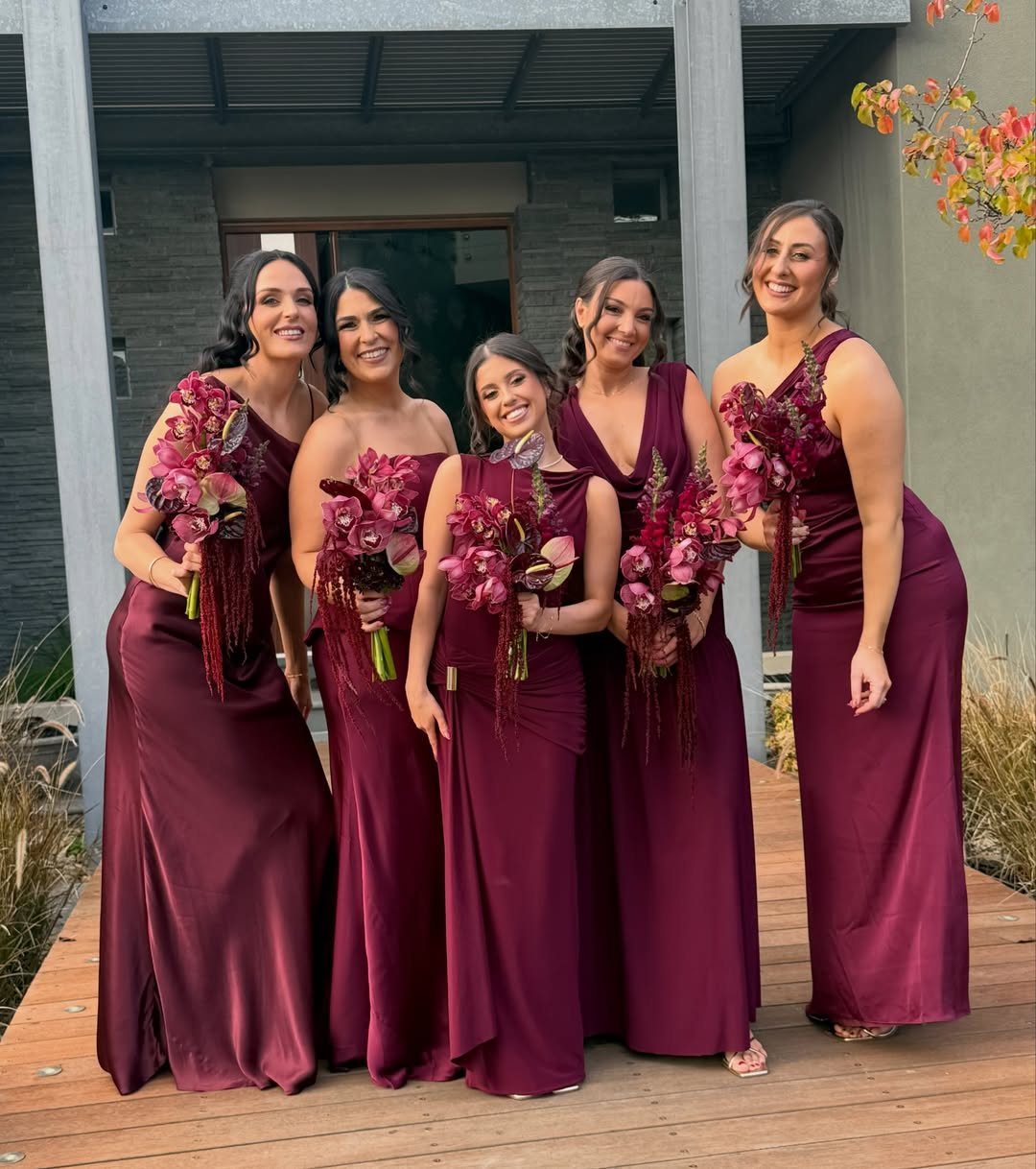 Five bridesmaids in burgundy dresses of different styles holding matching floral bouquets.