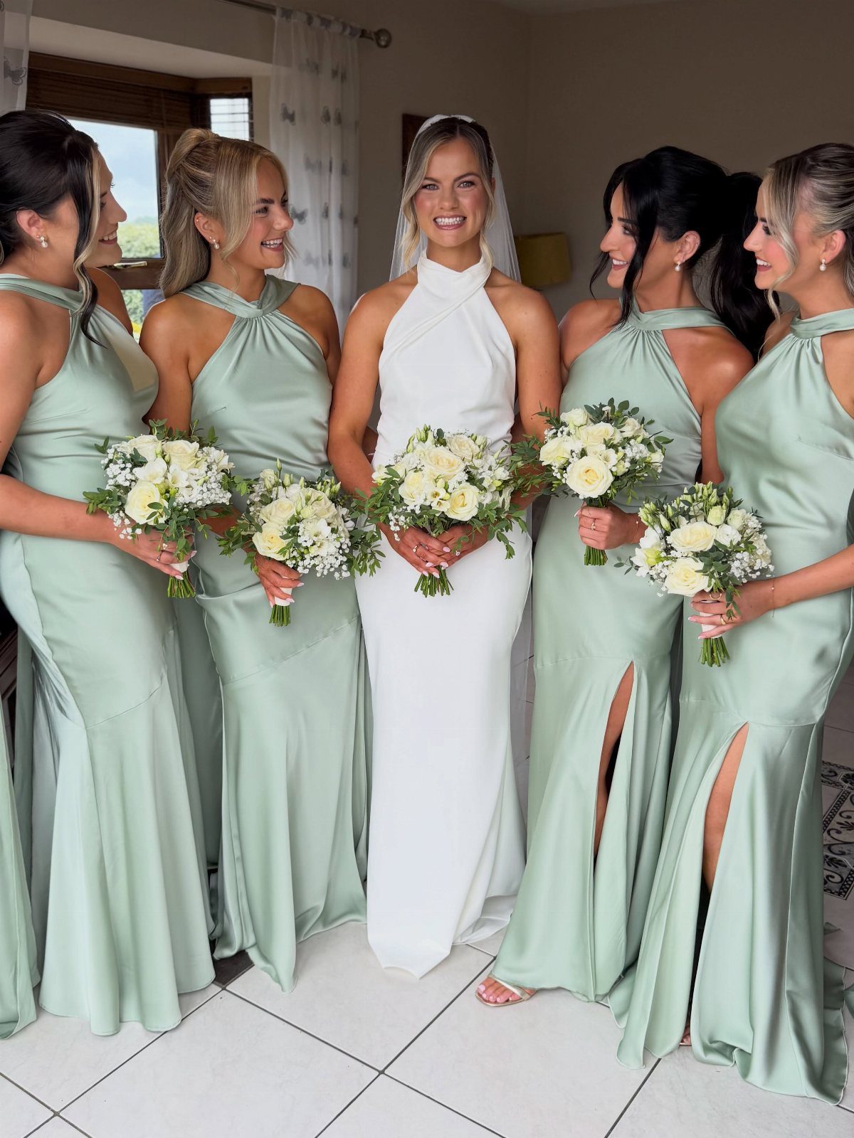 Bride in white gown with bridesmaids in sage green dresses holding white bouquets.