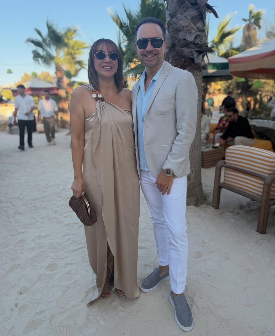 Couple in dressy beach attire posing on white sand with palm trees behind.