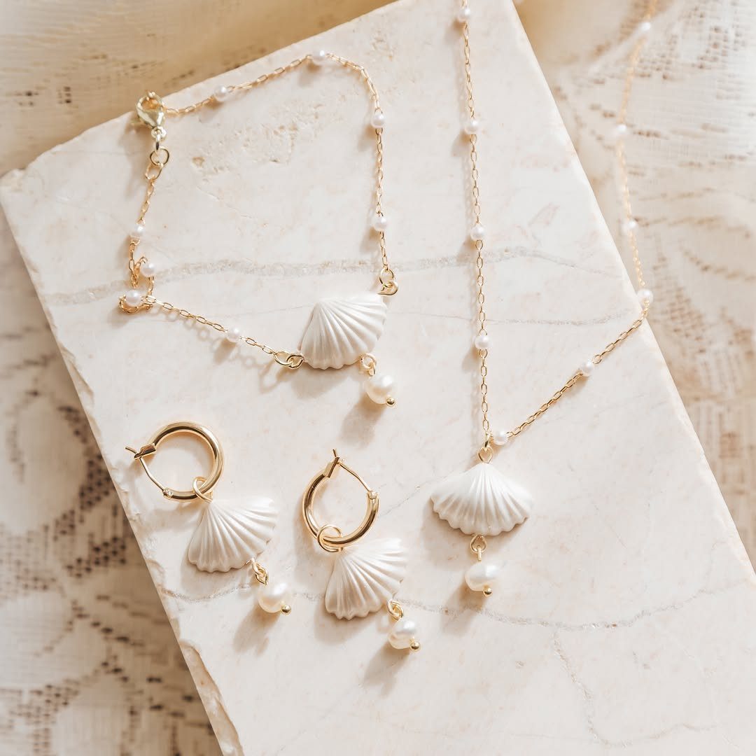 Gold jewelry set with white seashell charms and pearls on a stone slab.