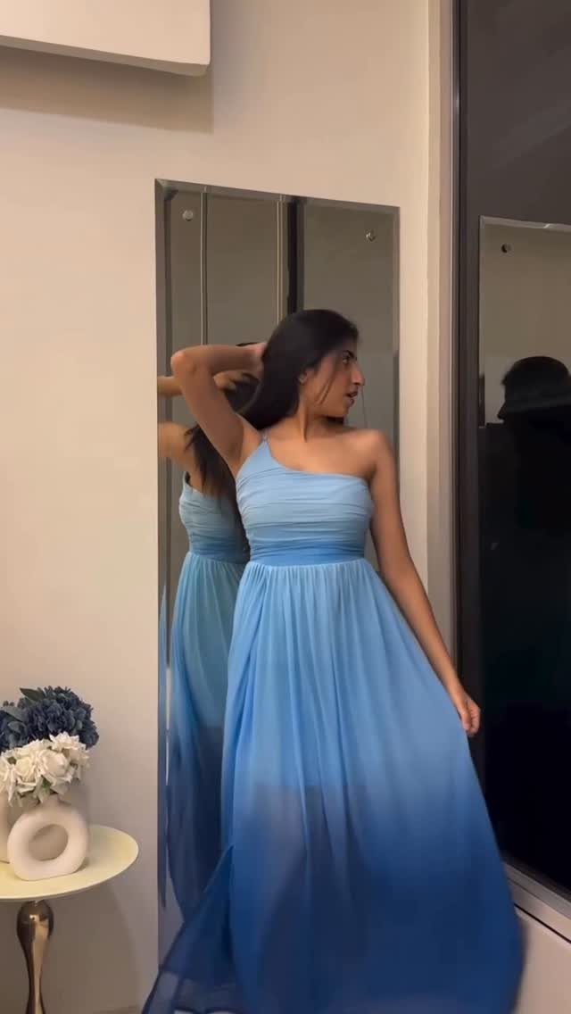 Woman wearing a light blue one-shoulder flowy maxi dress standing before a mirror.