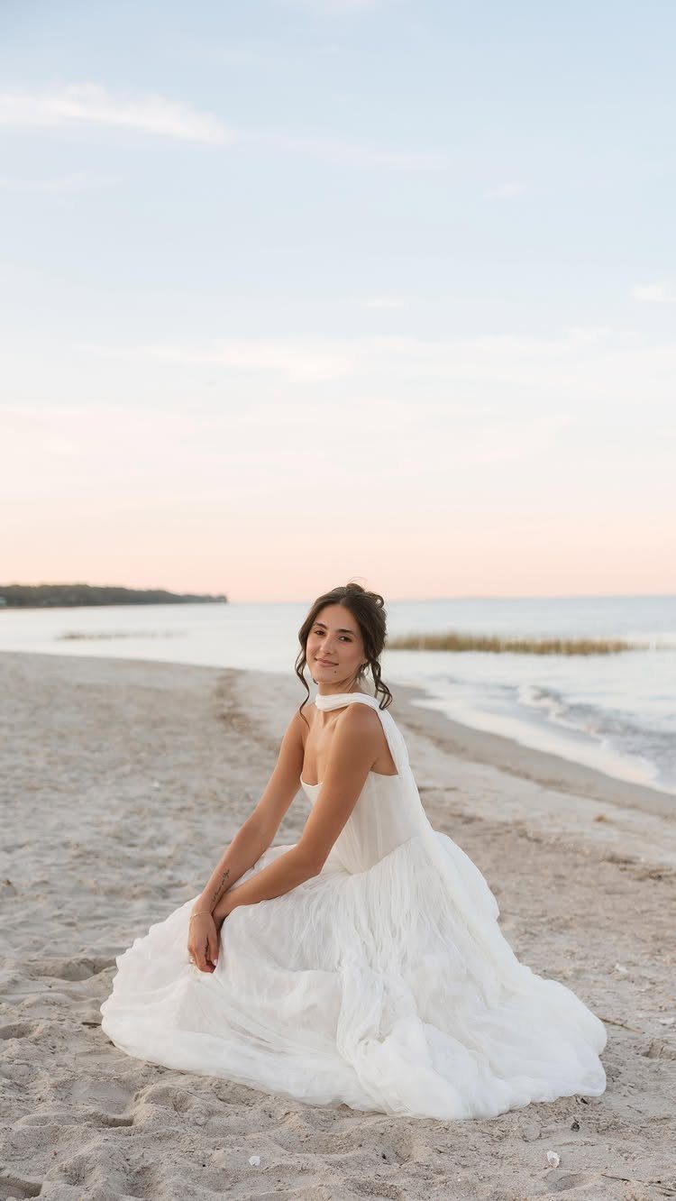 Woman in a flowy white wedding gown sitting on a sandy beach at sunset.
