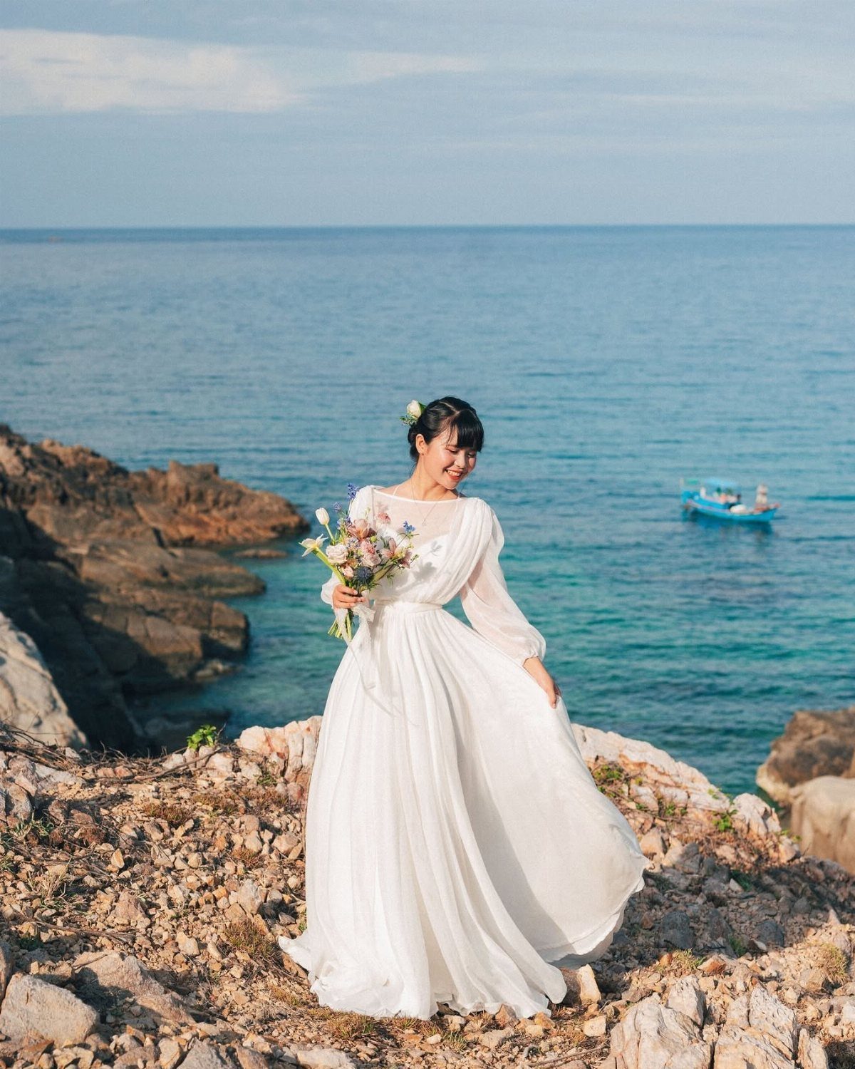 Woman in a white wedding gown holding flowers on a rocky ocean cliff