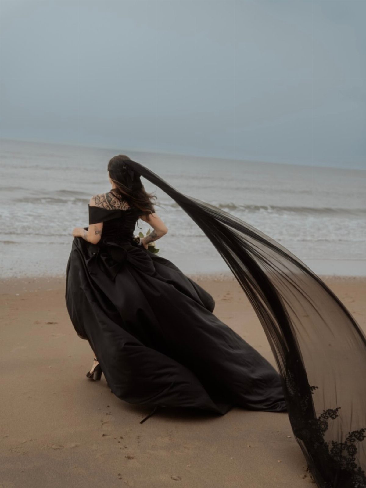 Woman in a voluminous black wedding dress and long veil on a sandy beach