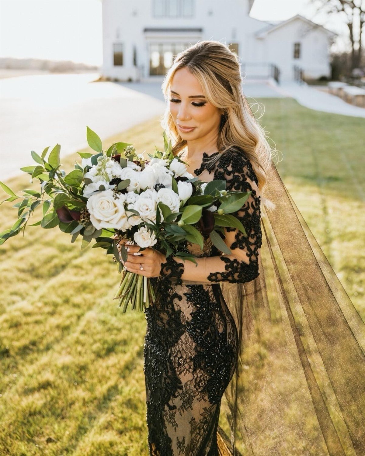 Woman in a form-fitting black lace wedding dress holding white rose bouquet outdoors