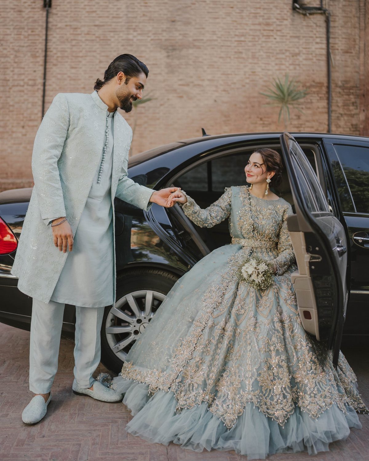 Couple in light blue traditional wedding attire standing beside a black car.