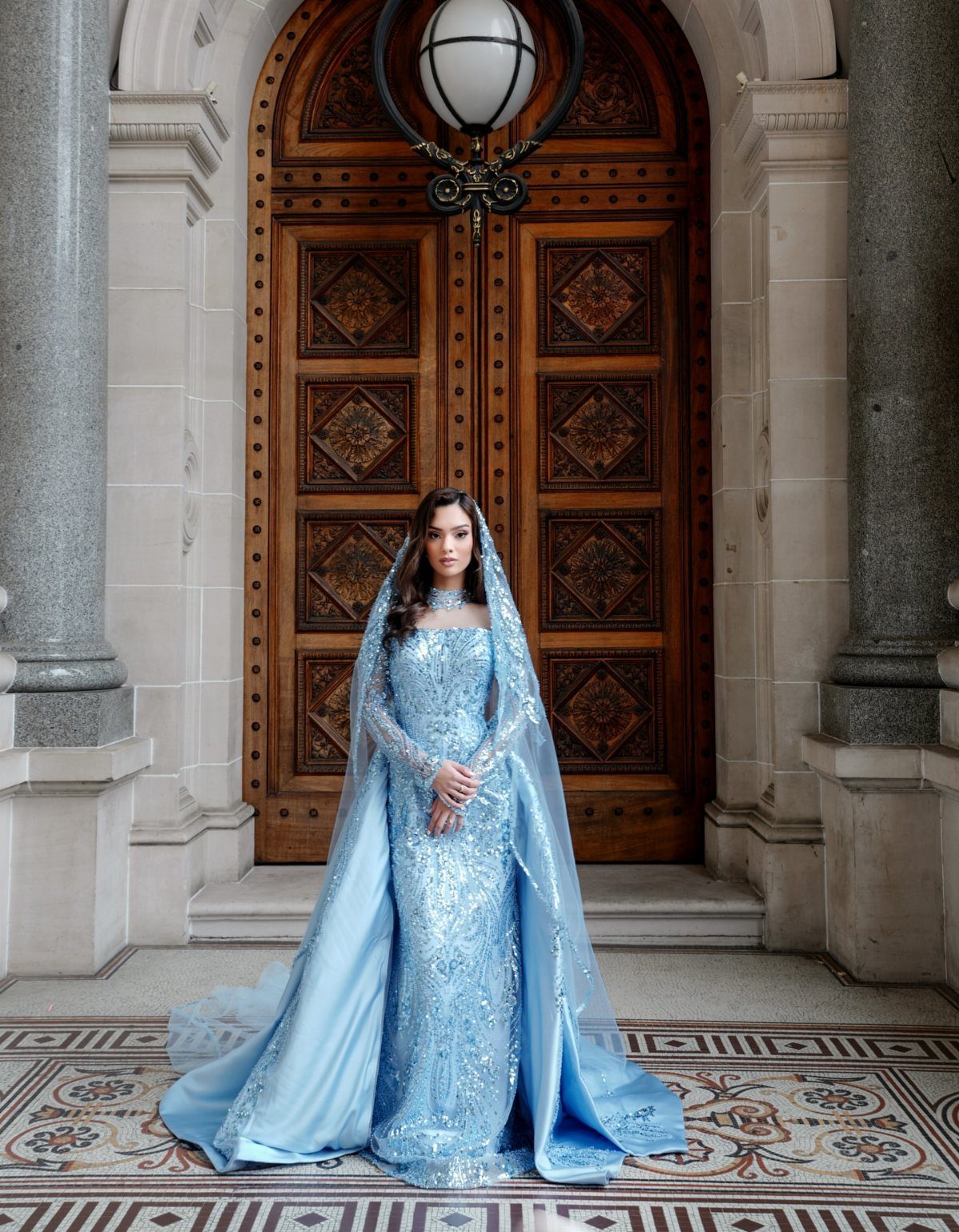 Woman in a shimmering light blue sequined bridal gown and veil before wooden doors