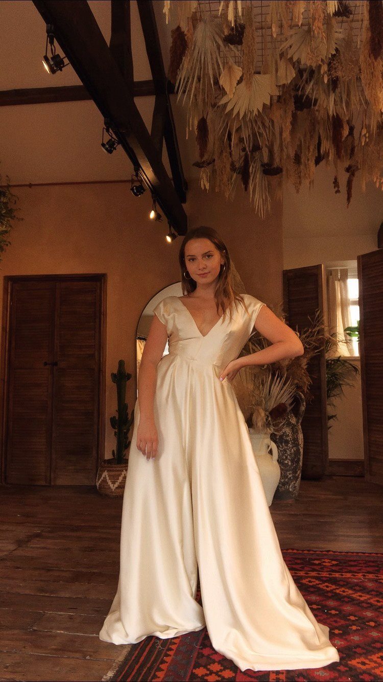 Woman in a wide-leg cream satin bridal jumpsuit standing in a rustic room.