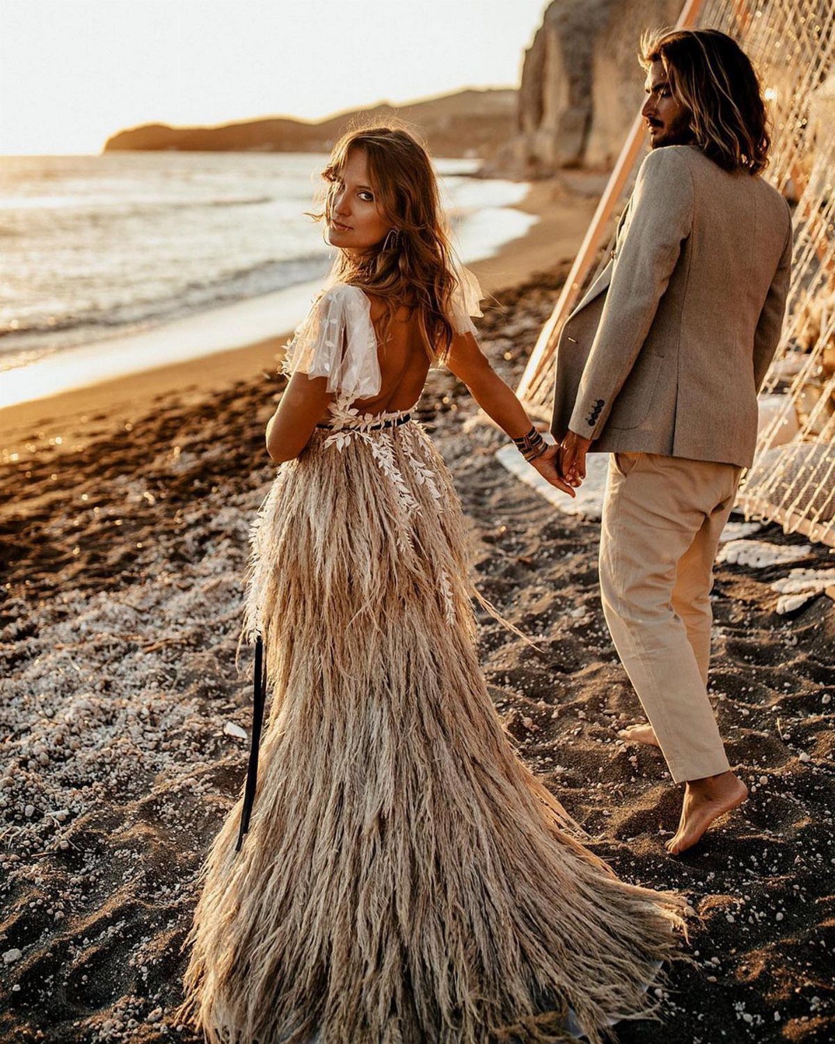 Woman in a fringed boho bridal gown walking on a beach with a man.