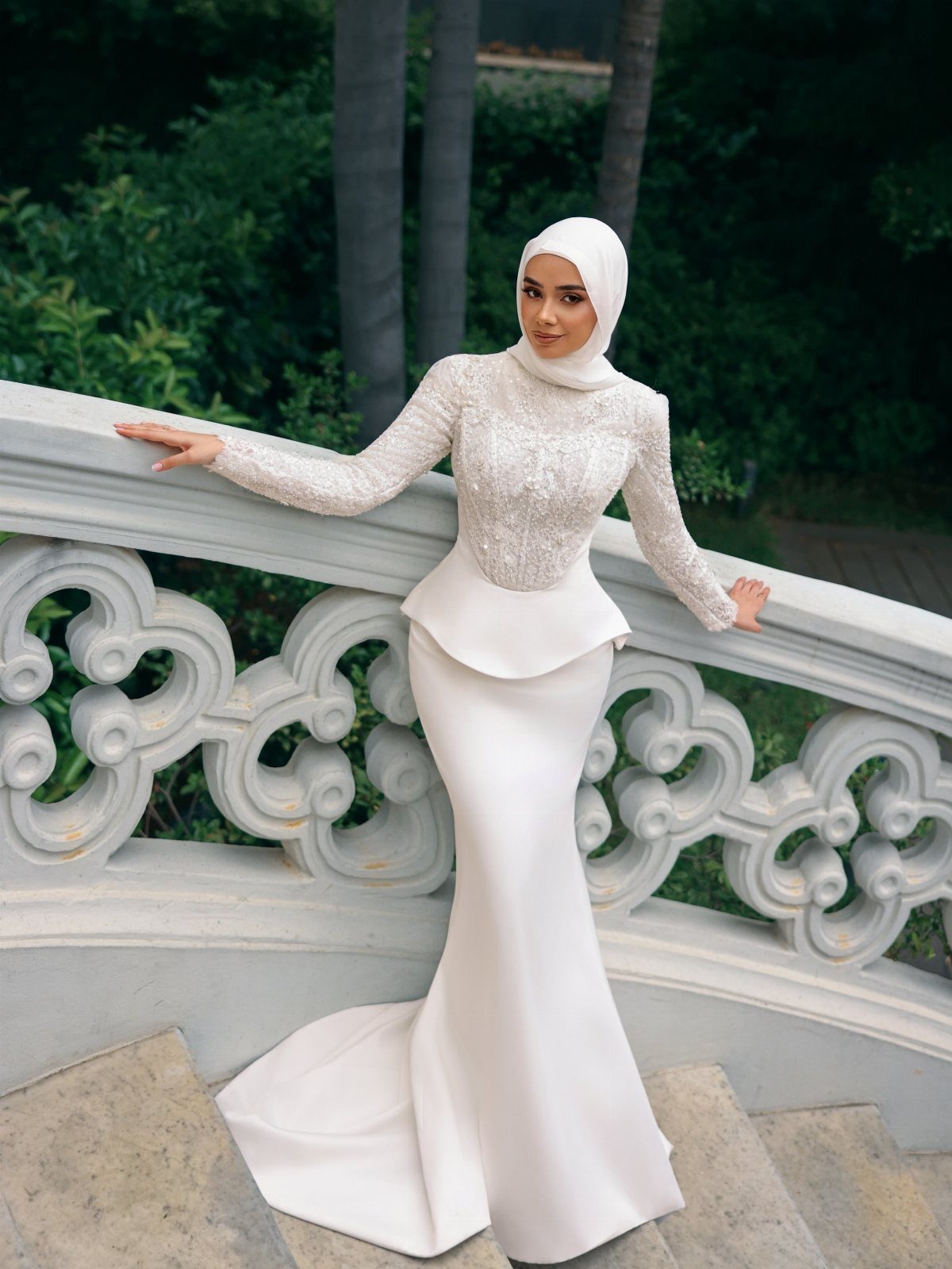 Woman in a white mermaid bridal gown and hijab posing by stone railing