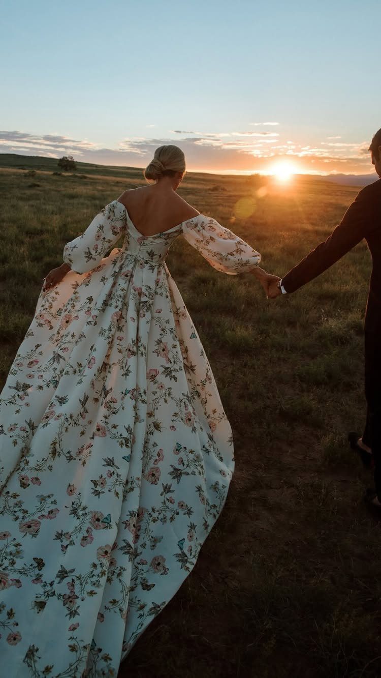Woman in floral wedding dress holding hands with man in a field at sunset