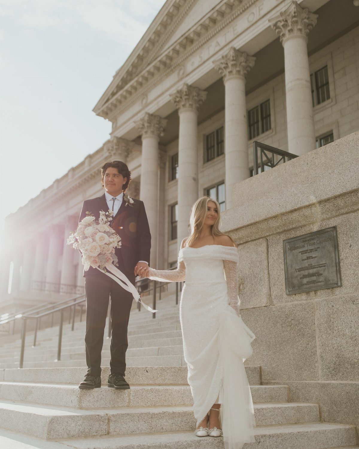 Bride in a white sheath wedding dress and groom in black suit holding hands.
