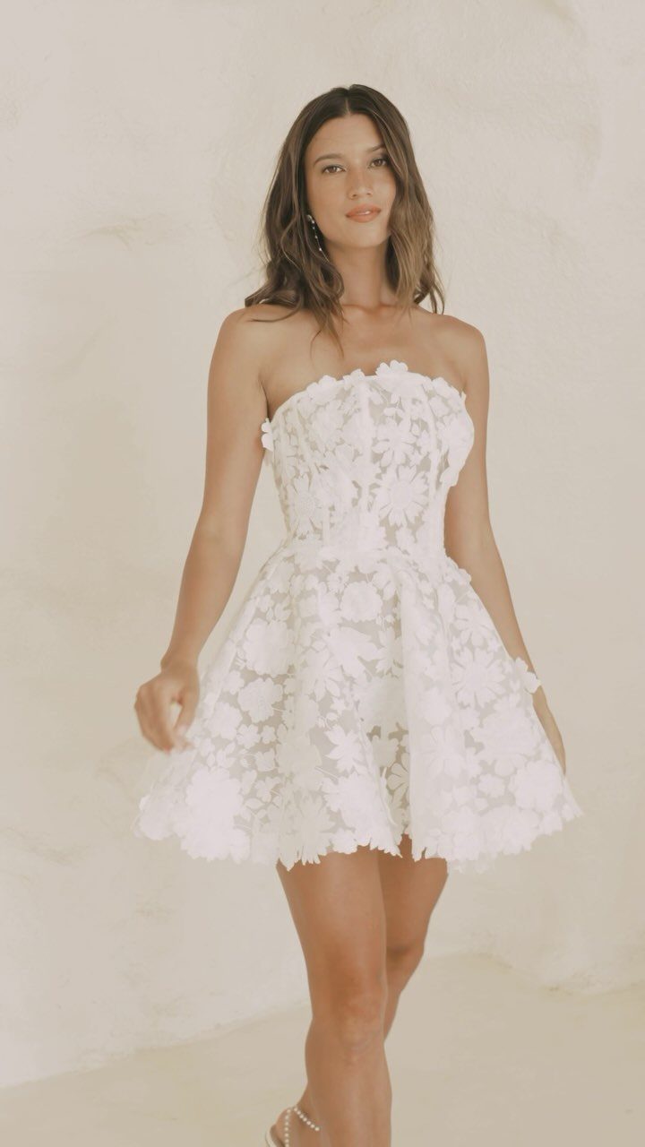 Woman wearing a short, strapless white floral lace wedding dress against a plain wall