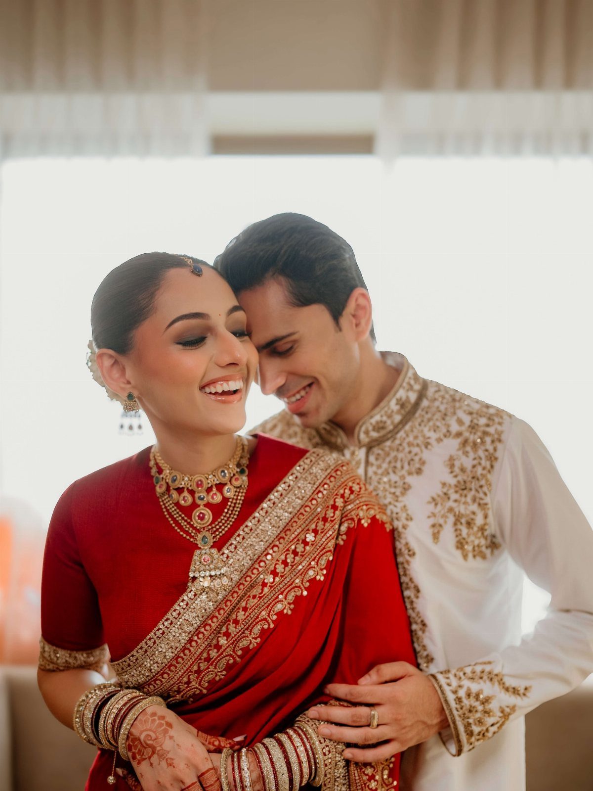 Smiling bride in red saree and groom in white sherwani embracing and laughing.