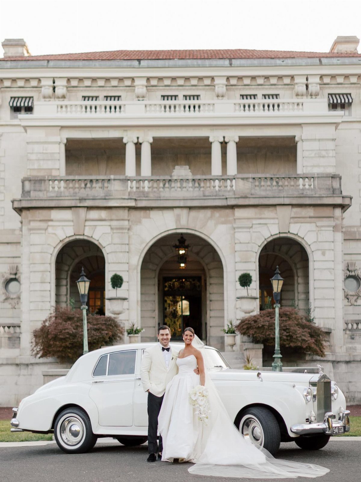 Bride and groom posing beside a vintage white luxury car before a mansion