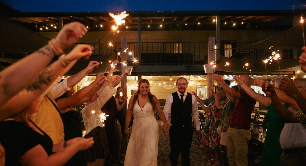Bride and groom walking through a tunnel of guests holding lit sparklers at night.
