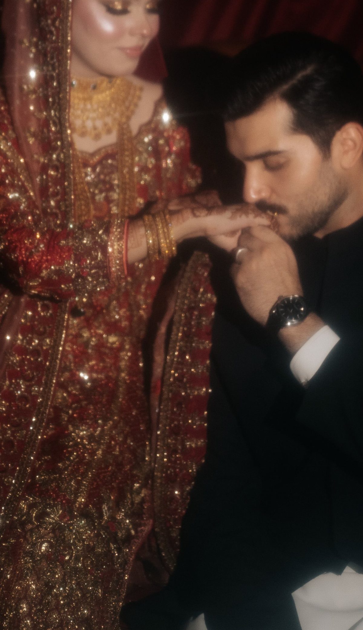 Groom in black suit kissing the hand of bride in red gold attire