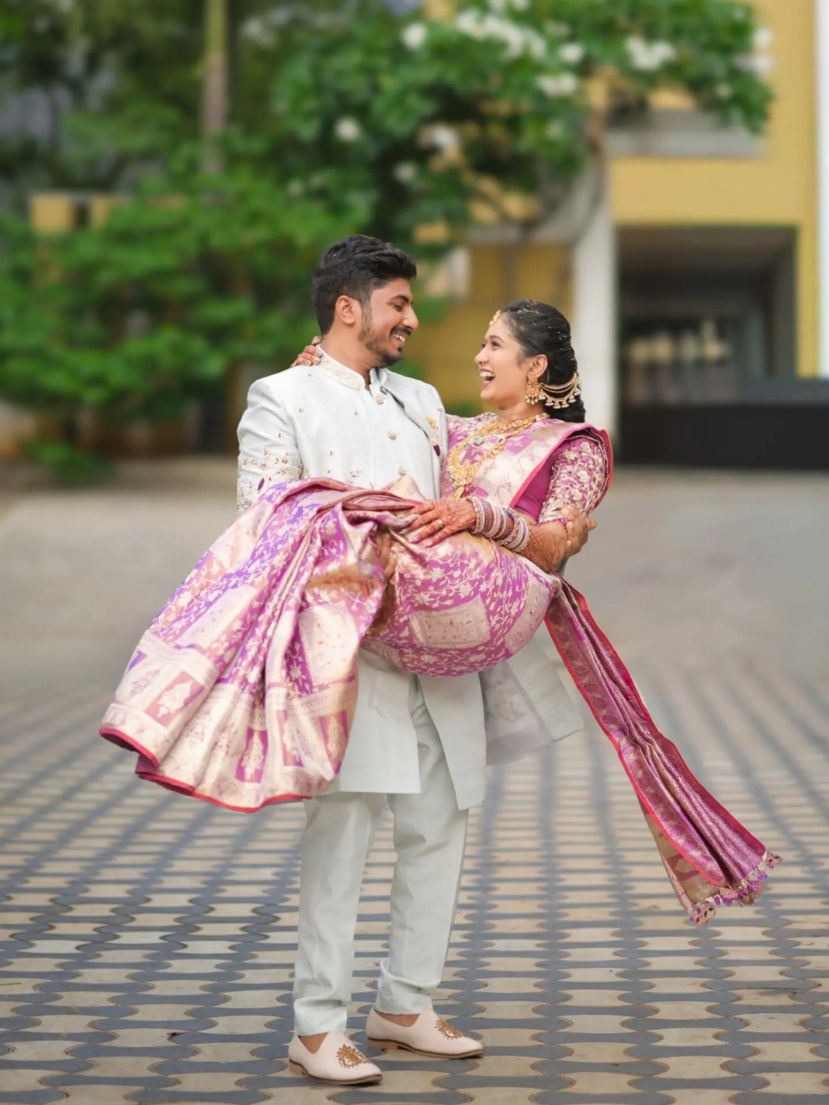 Groom in white sherwani lifting bride in purple silk saree outdoors