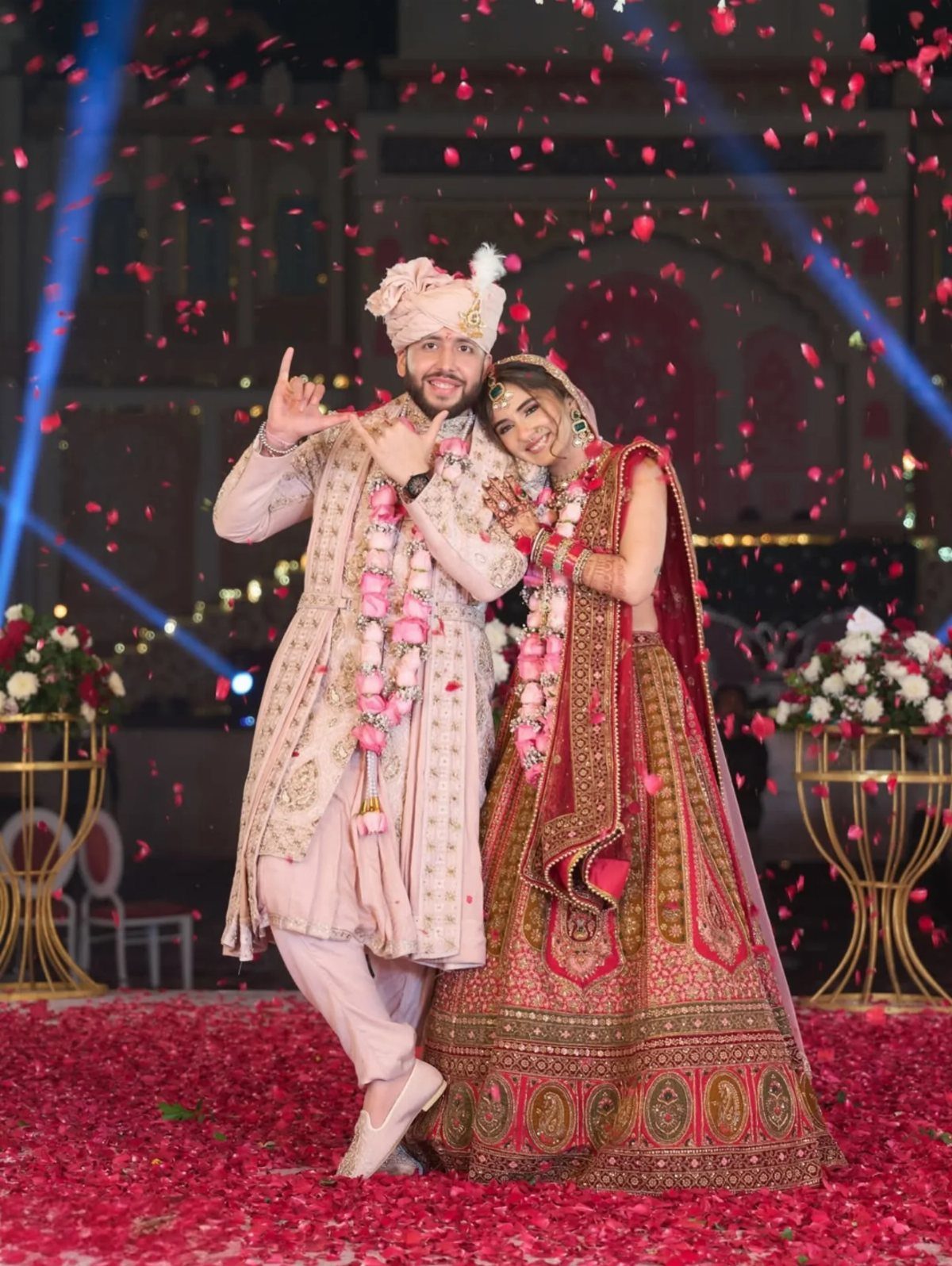 Bride and groom in traditional Indian wedding attire posing amidst falling red petals.