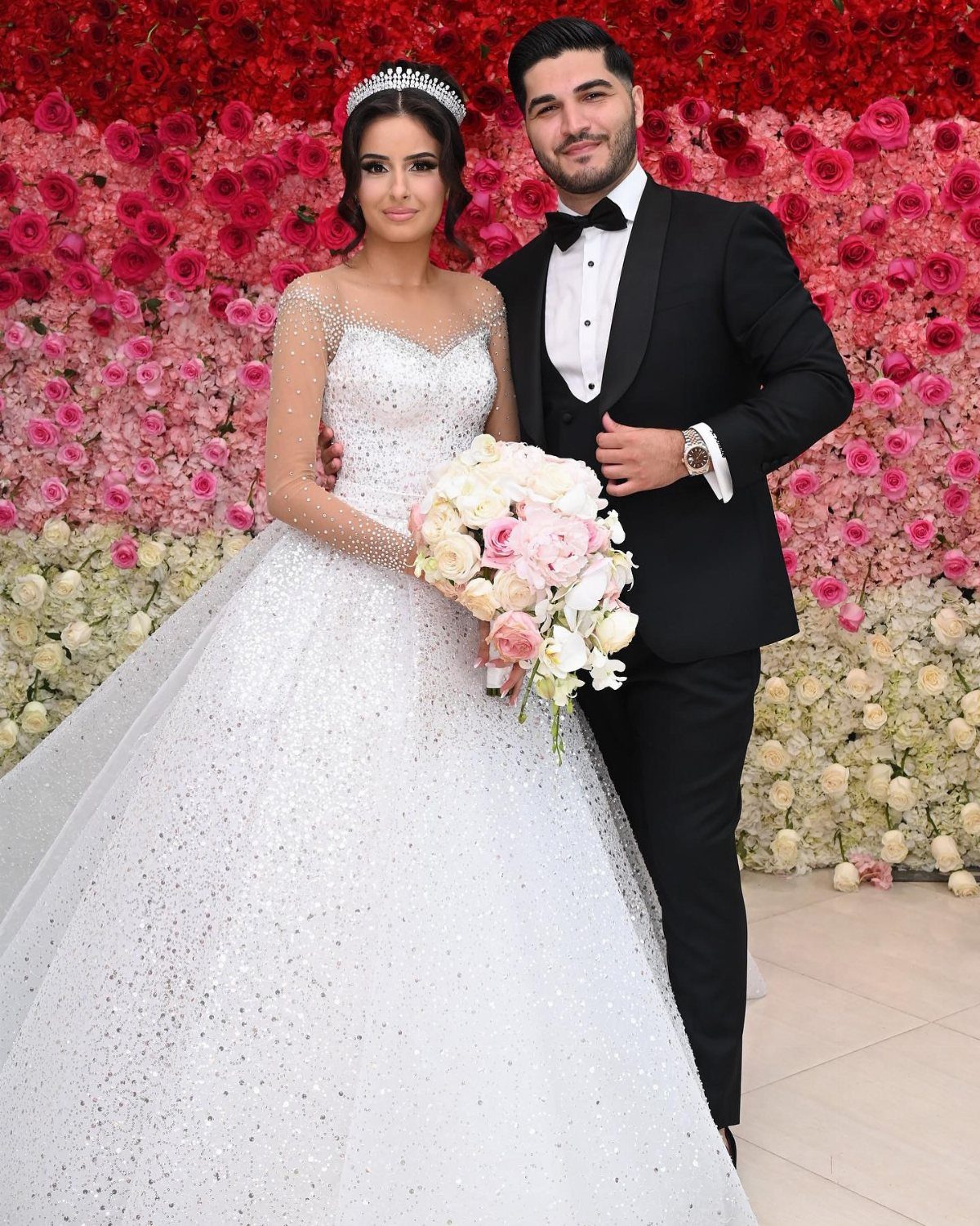 Bride in sparkling white gown and groom in black tuxedo posing before flower wall