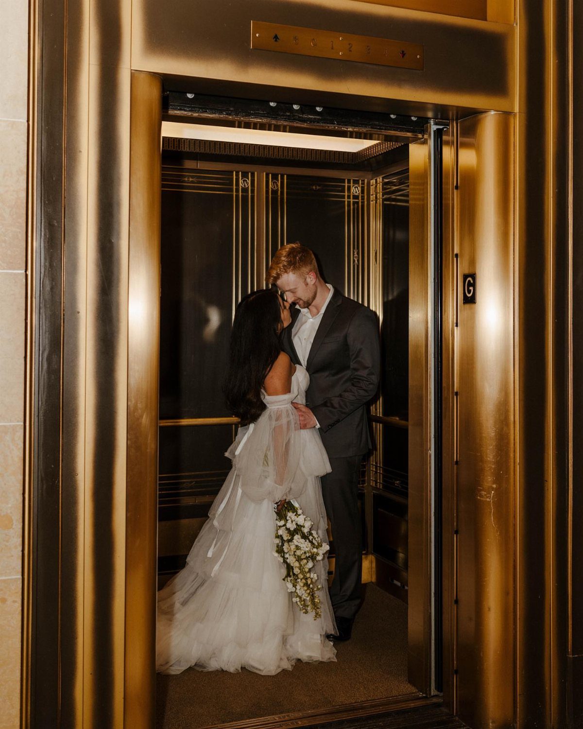 Bride and groom standing close together inside a gold elevator looking at each other