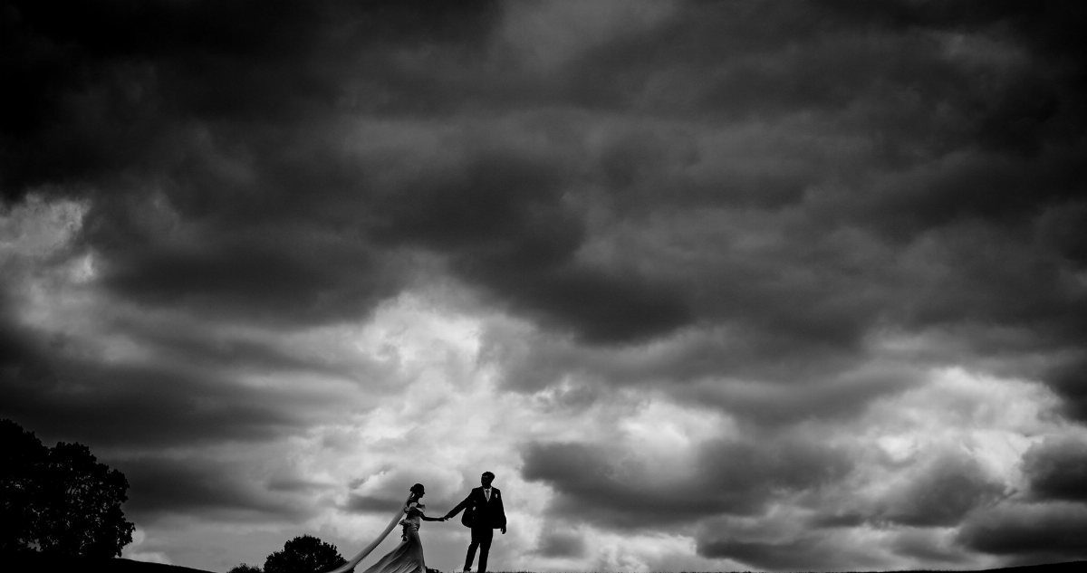 Bride and groom silhouettes holding hands under a dramatic, cloudy black and white sky