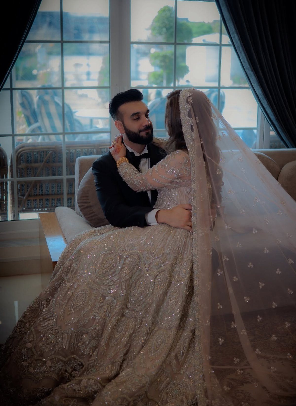 Bride in gold gown and groom in tuxedo sitting on a sofa together.