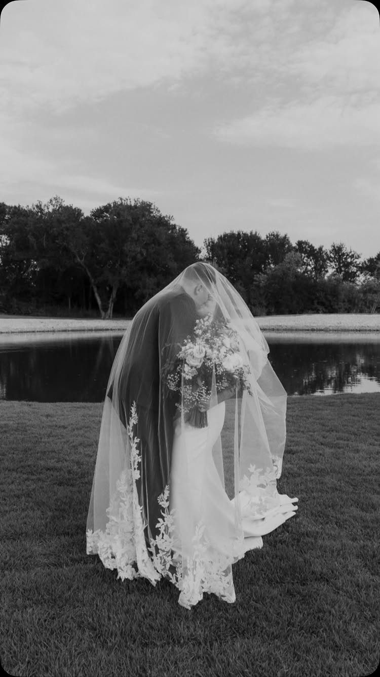 Bride and groom embracing under a long, lace-edged sheer wedding veil outdoors.
