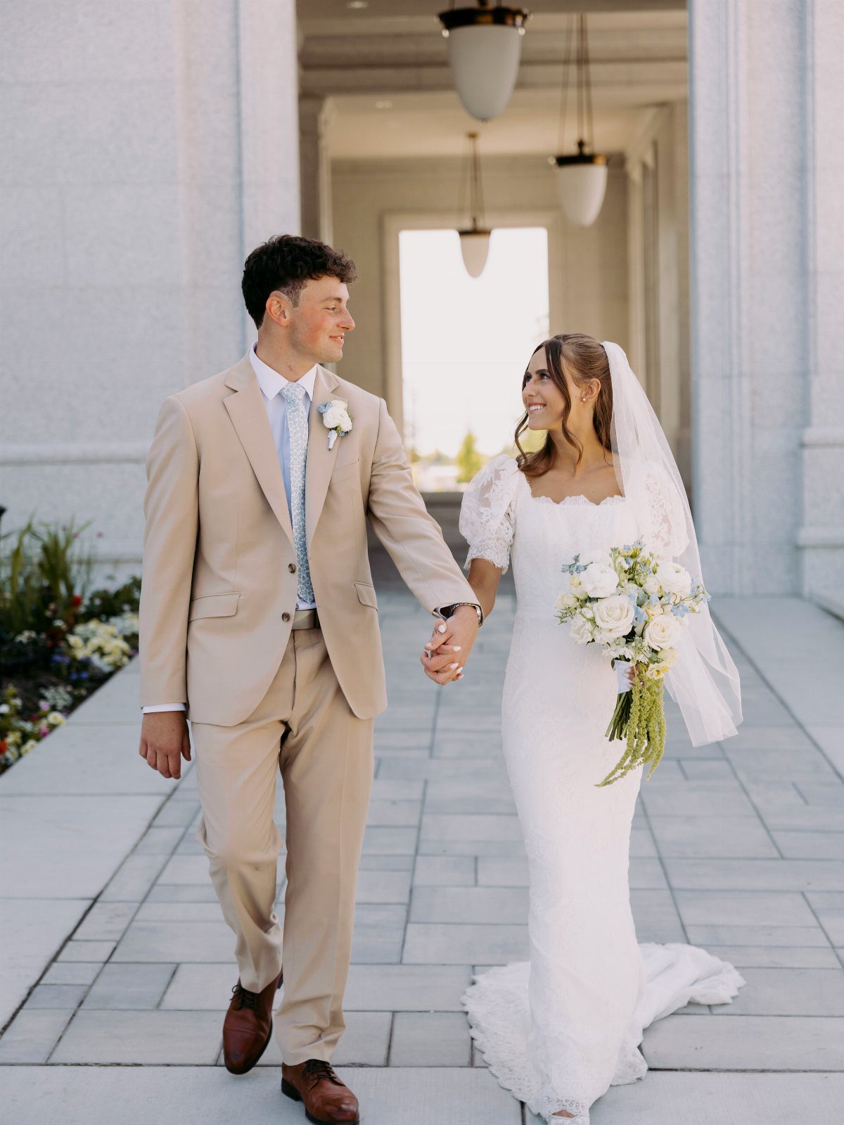 Bride and groom walking hand in hand on a stone path between pillars.