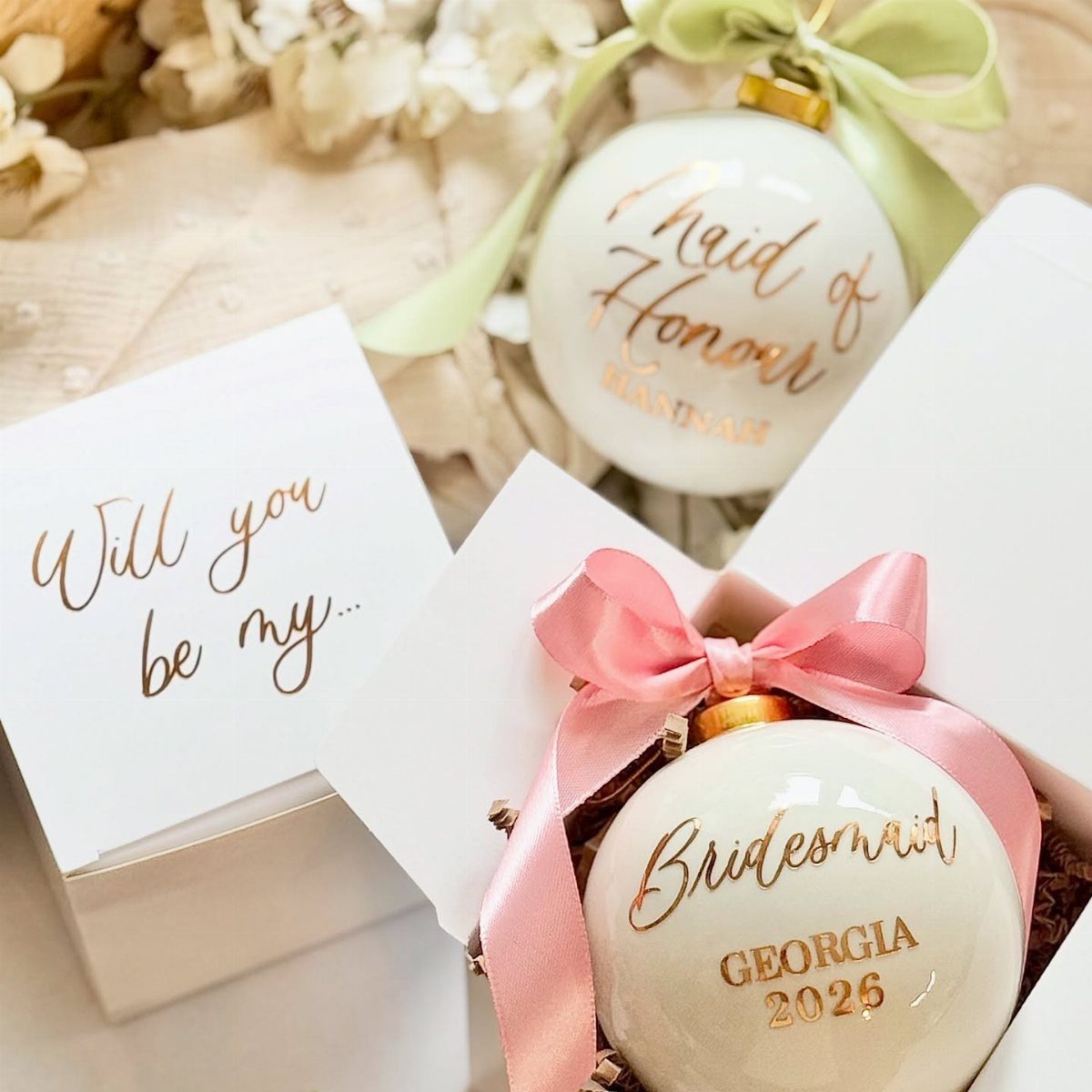 White Christmas ornaments with gold bridesmaid proposal text in white gift boxes.