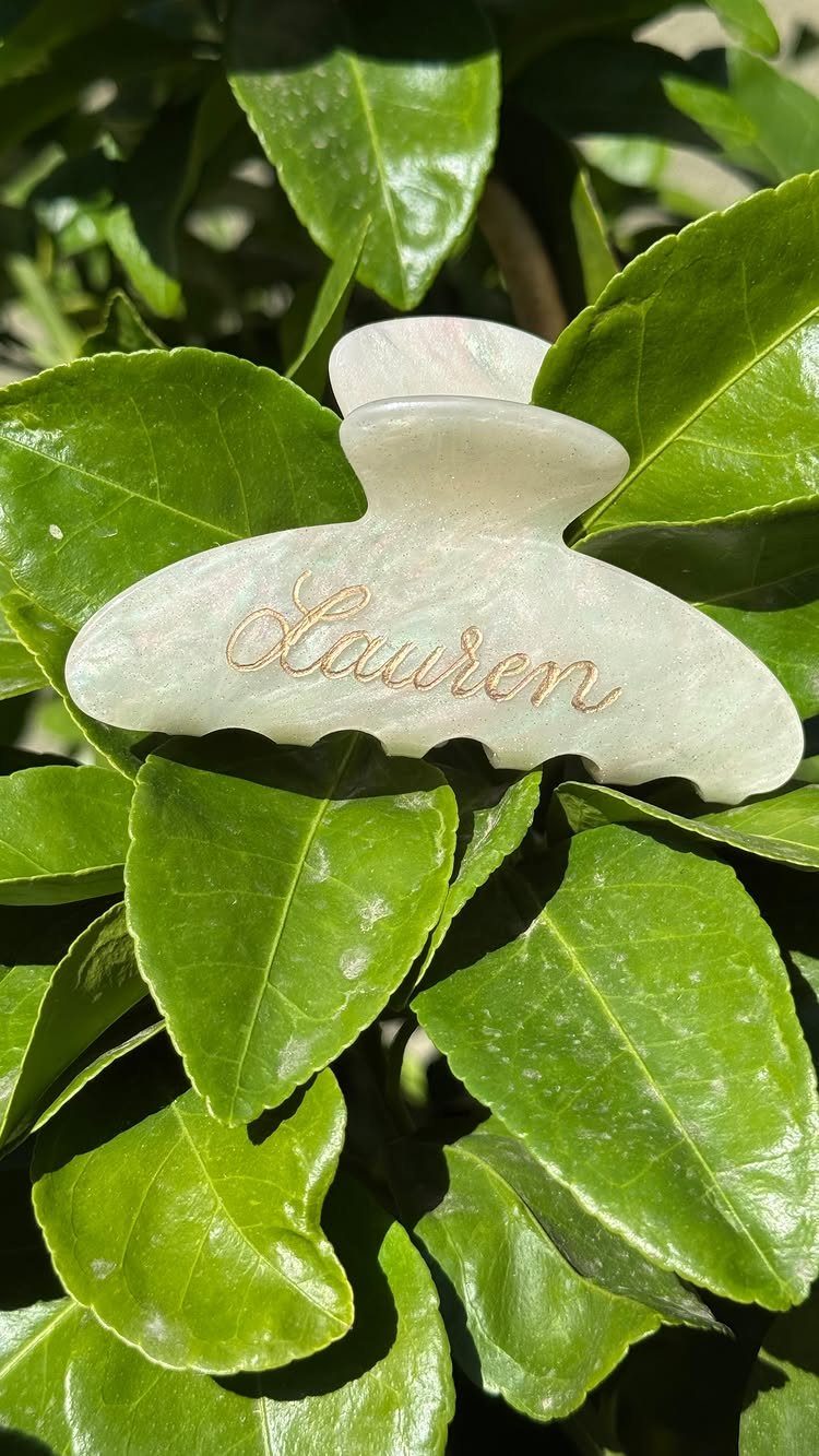 Pearlescent hair claw clip with gold name Lauren resting on green leaves