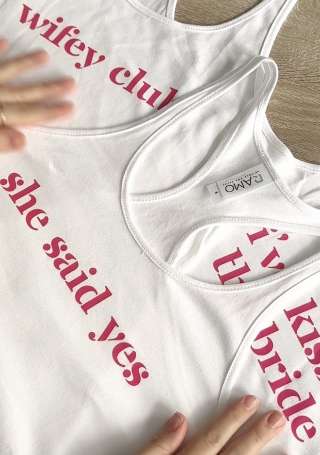 White tank tops with pink bridal phrases laid on a wooden table.