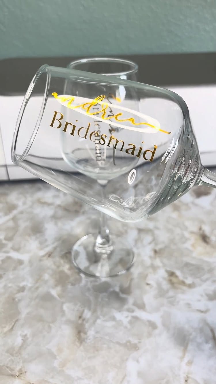 Two wine glasses with gold Bridesmaid lettering resting on a white marble countertop.