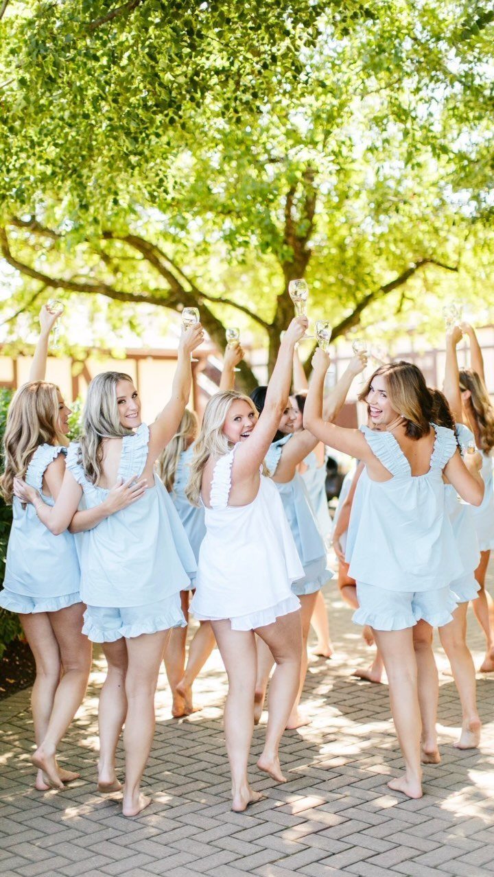 Bridesmaids in matching light blue and white pajamas cheering with champagne glasses outdoors.