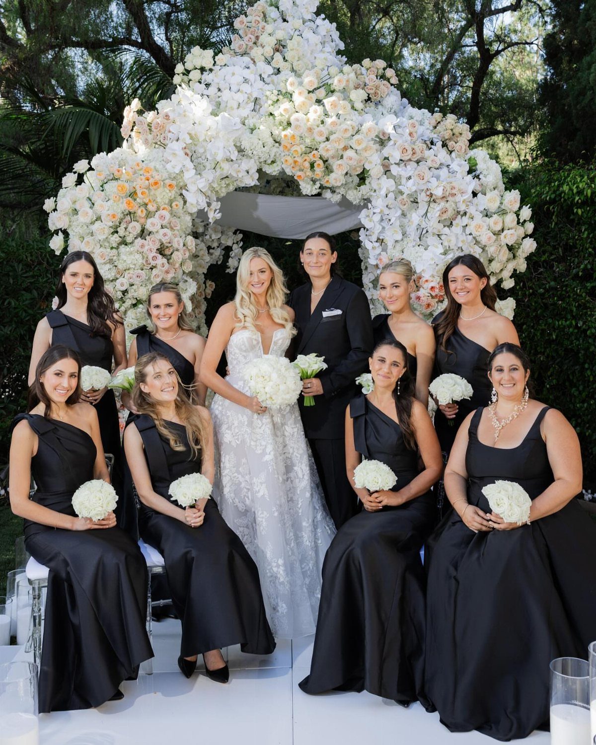 Bride and groom with bridesmaids wearing elegant long black dresses under a floral arch