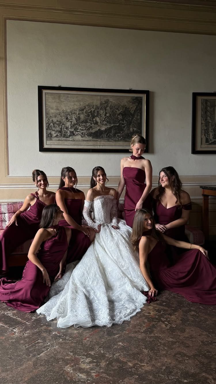 Bride in white lace gown with bridesmaids wearing matching burgundy dresses in a room.