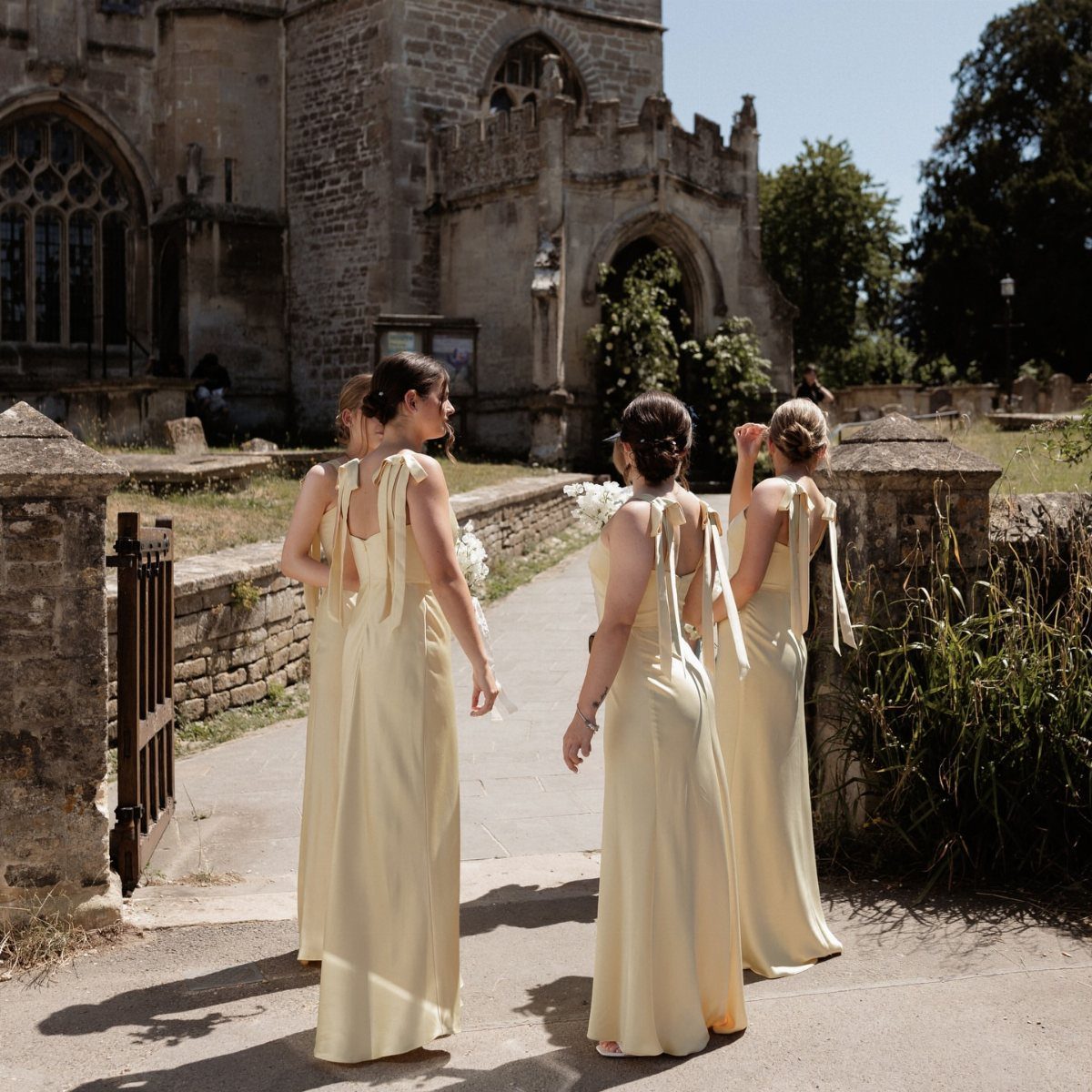 Four bridesmaids in pale yellow gowns walking toward an old stone church.