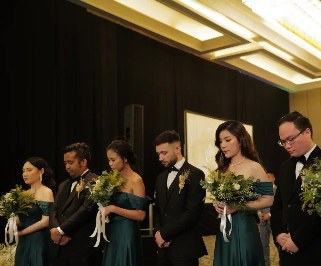 Bridesmaids in dark teal dresses and groomsmen in black tuxedos at a wedding.