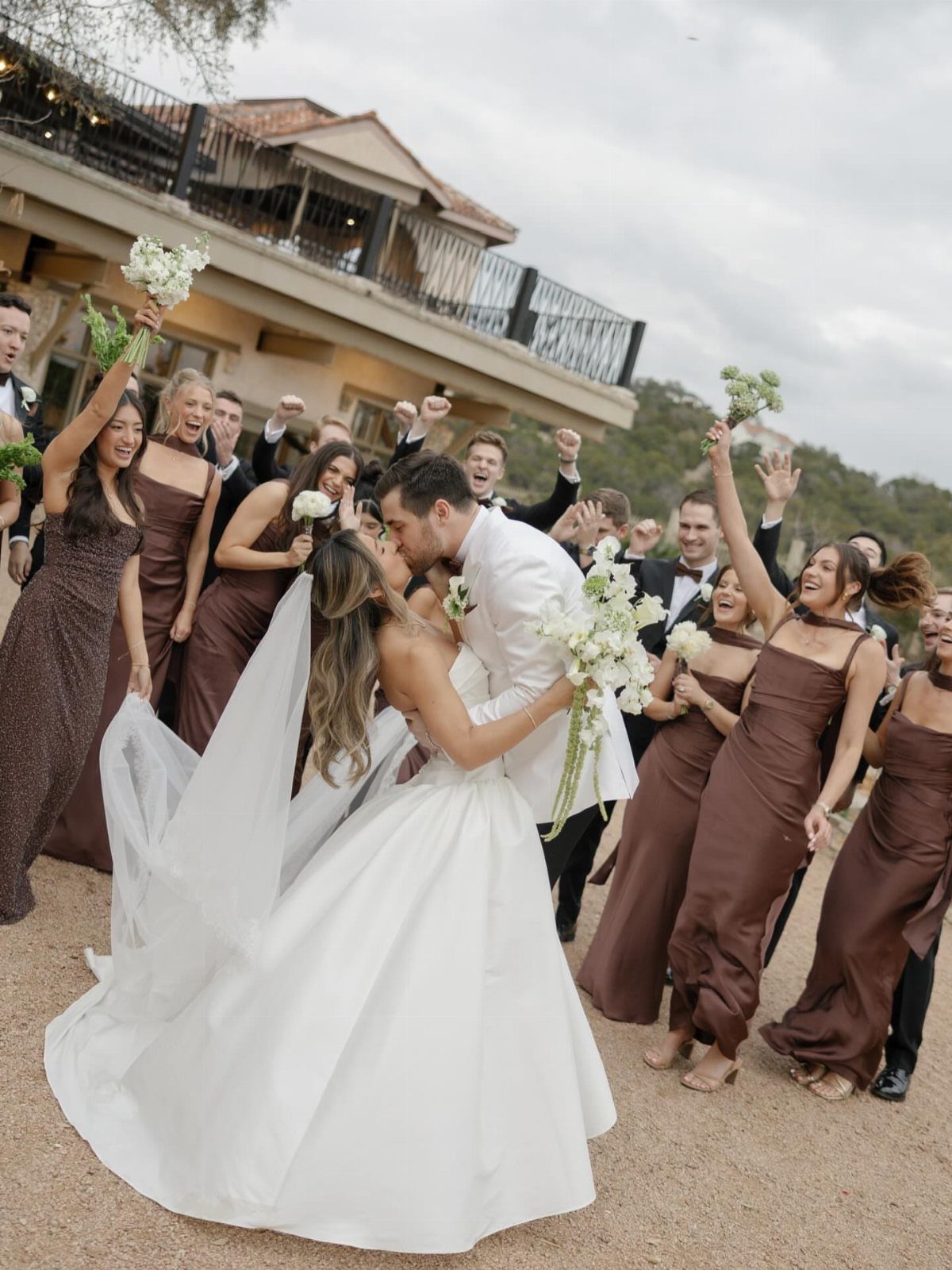 Bride and groom kissing surrounded by bridesmaids in brown dresses and groomsmen.
