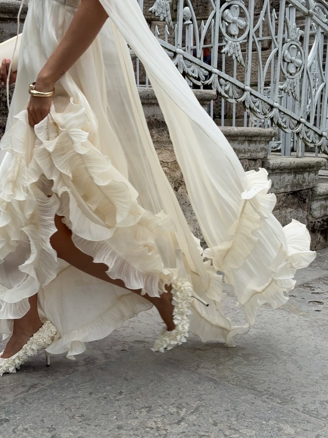 Cream wedding dress with voluminous ruffles and a matching ruffled cape flowing behind.